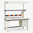 Industrial workbench with beige metal frame, white laminate surface, overhead fluorescent light, upper shelf, and colorful red, yellow, and blue storage bins. Includes built-in power strip and two-drawer side cabinet, set against a seamless white background.