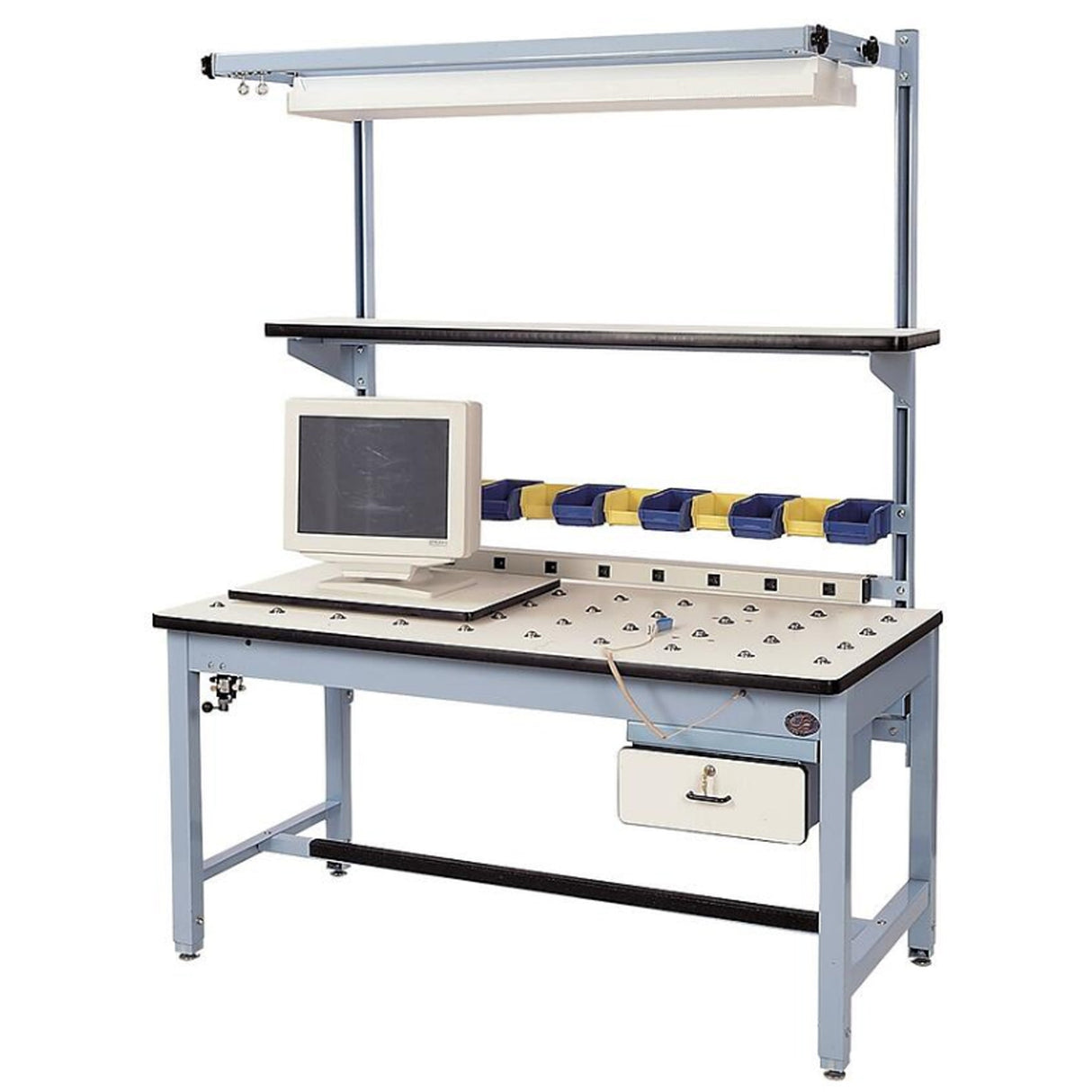 Rotating-top industrial workstation with a white perforated surface, mounted CRT monitor, multiple small parts bins in yellow and blue, overhead lighting, upper utility shelf, and integrated power strip on a light blue steel frame.